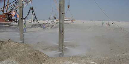 Eine große Baugrube ist eingehüllt in eine Sandwolke; Arbeiter sind zu sehen, die den Baugrund bearbeiten.