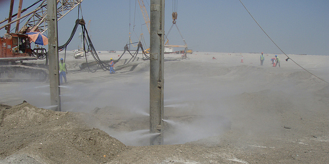 Eine große Baugrube ist eingehüllt in eine Sandwolke; Arbeiter sind zu sehen, die den Baugrund bearbeiten.