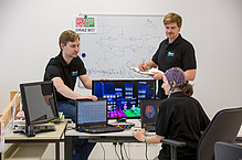 Woman with wired hood in front of several screens with brain models, behind her two men.