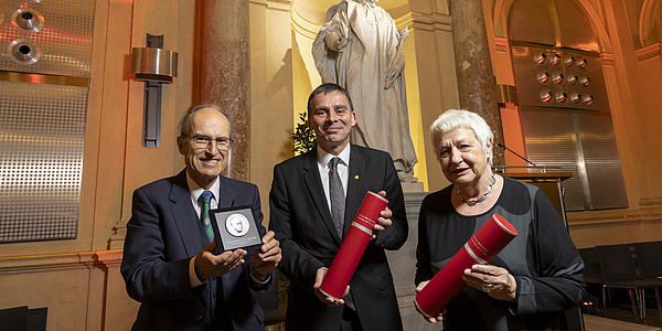 Eine Frau und zwei Männer, zwei mit Urkundenrollen in den Händen, einer mit einer Medaille in einer Schatulle.