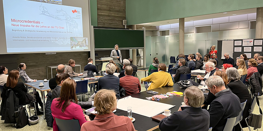 Viele Menschen sitzen an Tischen in einem Hörsaal. Ein Mann steht vor ihnen und hält einen Vortrag zum Thema Microcredentials.