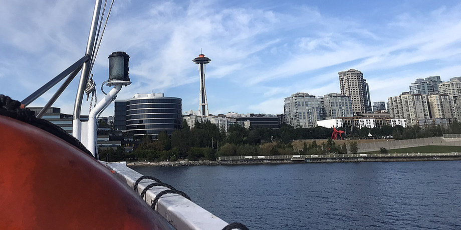 The skyline of a city, in front of it the sea, on the left in front the bow of a boat.