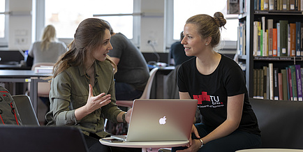 Zwei Frauen im Gespräch, zwischen ihnen ein aufgeklappter Laptop.