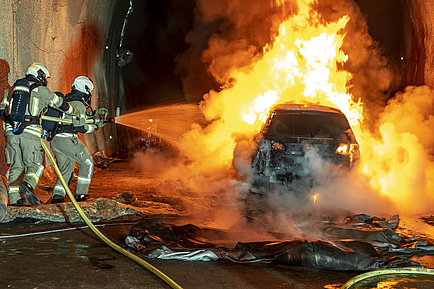 Zwei Feuerwehrleute spritzen Wasser auf ein brennendes Auto.