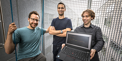 Moritz Lipp, Michael Schwarz und Daniel Gruss stehen in einem Durchgang. Daniel Gruss hält einen Computer, auf dem weißer Text auf schwarzem Hintergrund zu sehen ist.