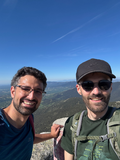 Two men smile at the camera during a sunny mountain hike, with a wide valley and clear blue sky in the background.