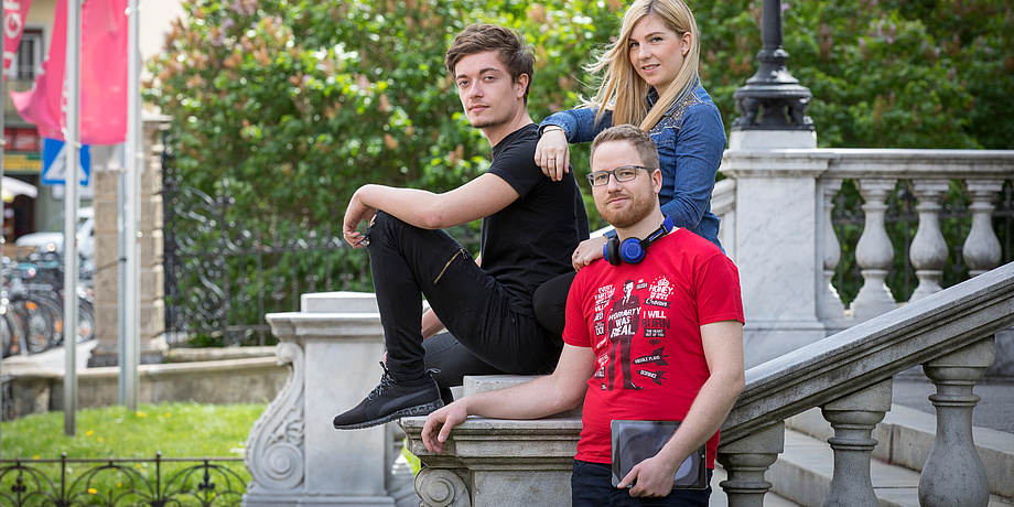 Two male and a female student at the picturesque staircase in front of the TU Graz old campus, with leafy trees in the background.