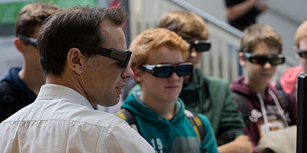 A TU Graz researcher surrounded by pupils at the Geoday. All wear VR goggles while looking at a screen which is hardly to be seen on the edge of the image.