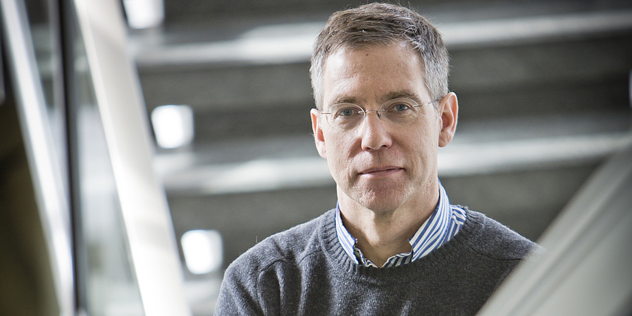 A man stands in a staircase. He wears a grey jumper and a shirt.