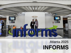 Zwei Personen posieren hinter einem großen blauen „INFORMS“-Schriftzug mit grünem Pfeil im Georgia World Congress Center in Atlanta 2025, flankiert von Bildschirmen in einer modernen Halle.