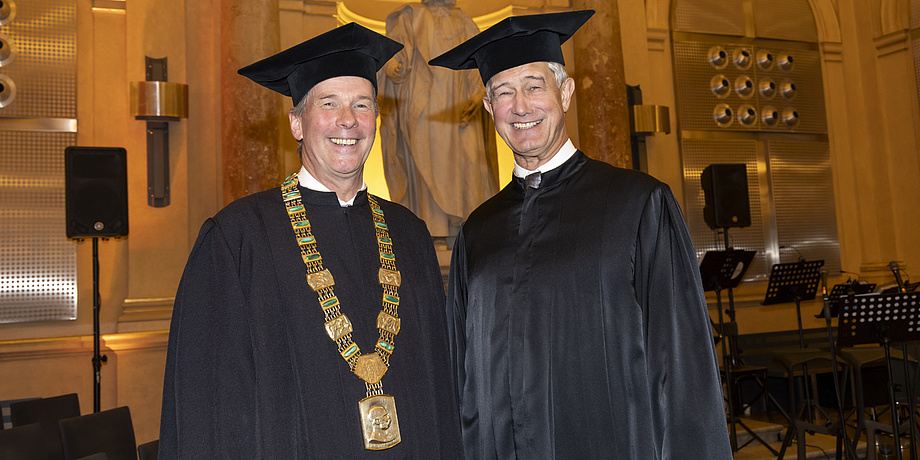 Two men dressed in robes stand next to each other and look into the camera.