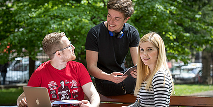 Zwei Burschen und ein Mädchen -  alle Studierende der TU Graz - unterhalten sich im Park miteinander. Vor ihnen ein Laptop.