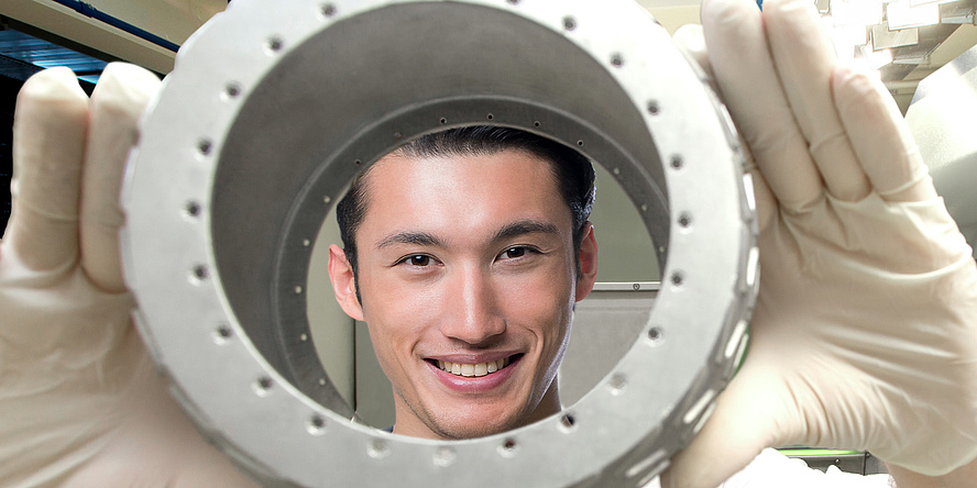 A friendly smiling young man looks through a round metal object into the camera, the TU Graz logo can be seen on the white polo shirt.