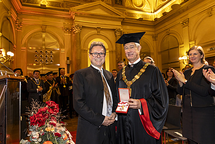 Two men smile into the camera, the man on the right wears a academic hat and holds a decoration of honor in his hand