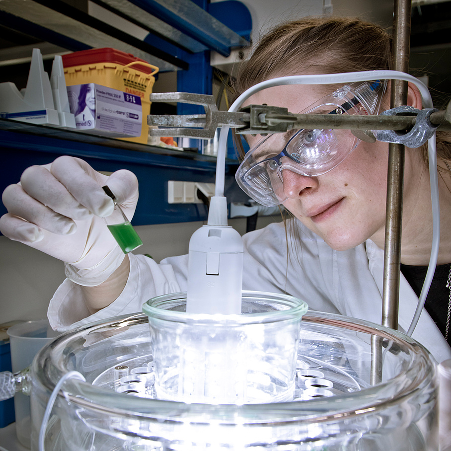 Woman with protective goggles bends over an illuminated glass container.