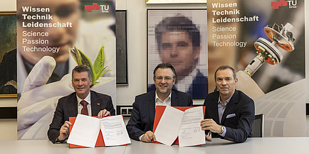 Three men sit next to each other and hold up two signed contract documents.