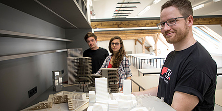 Zwei Studenten und eine Studentin zeigen stolz Architekturmodellen in den Architekturstudios der TU Graz.