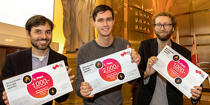 Three men joyfully hold a certificate informing of an award into the camera. The man on the left wears a dark beard, the man on the right wears glasses and a beard.
