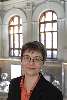 A woman in an orange blouse smiles, standing in front of two large windows.