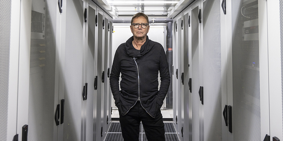 A man with short hair, glasses and black clothes stands between grey network cabinets.
