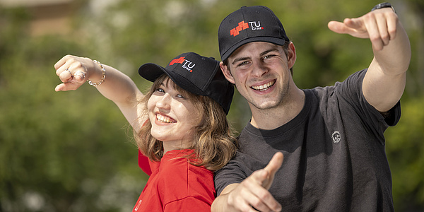 Eine junge Frau mit rotem T-Shirt und ein junger Mann mit schwarzem T-Shirt lachen in die Kamera. Beide tragen Schirmmützen mit TU Graz Logo.