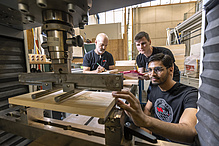 Drei Männer stehen hinter einer Maschine, in die ein Stück Holz eingespannt ist.