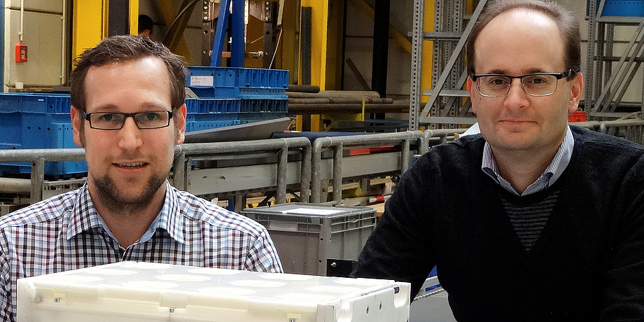 TU Graz researchers Florian Ehrentraut and Christian Landschützer (with black pullover over a small checkered shirt) proudly demonstrate the white prototype of a modular transport box from the MODULUSHCA research project in a lab at the Institute of Logi