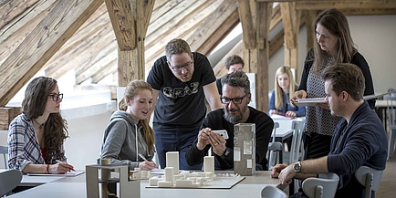Studierende und ein Lehrender diskutieren über ein Architekturmodell, das auf einem Tisch steht.