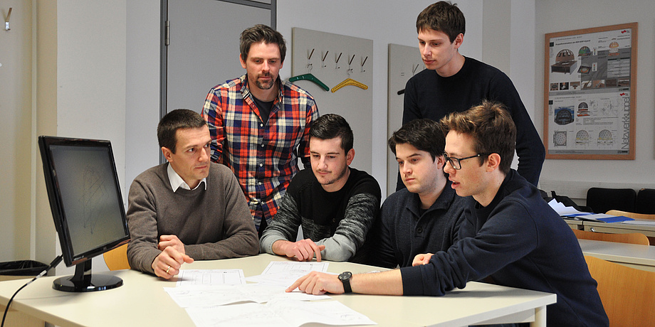 Four students and two supervisors work in front of a computer on a white table with work documents on the project for the steel bridge competition.