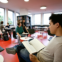 Ein Mann sitzt in einem Aufenthaltsraum und liest in einem Buch. Im Hintergrund mehrere Menschen. Bildquelle: Lunghammer – TU Graz