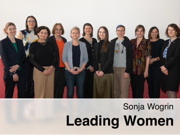 Eine Gruppe von zwölf Frauen steht in einer Reihe vor einer weißen Wand und einem roten perforierten Paneel, lächelnd in die Kamera, mit dem eingeblendeten Text „Sonja Wogrin – Leading Women“.