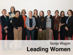 Eine Gruppe von zwölf Frauen steht in einer Reihe vor einer weißen Wand und einem roten perforierten Paneel, lächelnd in die Kamera, mit dem eingeblendeten Text „Sonja Wogrin – Leading Women“.