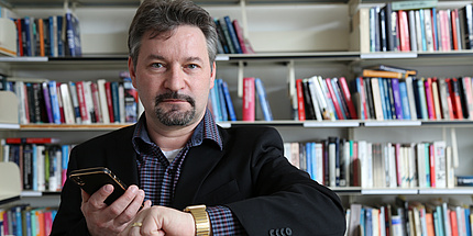 A man is standing in front of a bookshelf. He has raised both hands to chest level. He is holding a smartphone in one hand and a Smartwatch in the other.