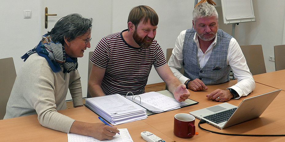 Zwei Männer und eine Frau sitzen am Tisch und schauen in einen Laptop. Vor ihnen befinden sich verschiedene Unterlagen.