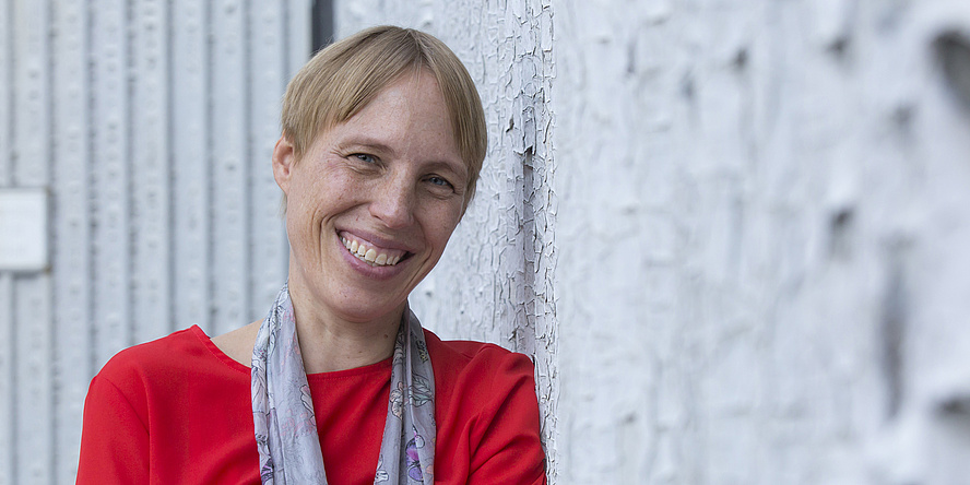 A woman with short blond hair leans against a gray wall.