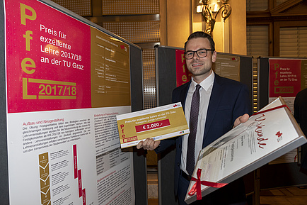 A man is holding a tribute in his right and left hand. To the left of him is a poster wall showing a poster in A1 format.