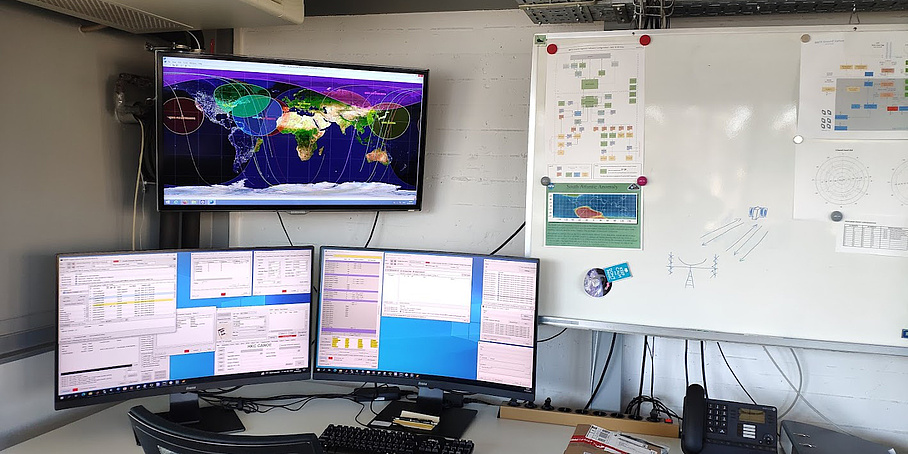 Ein Schreibtisch, auf dem zwei Computermonitore stehen, auf denen Daten zu sehen sind. Darüber hängt ein dritter Monitor an der Wand, auf dem Satellitenflugbahnen abgebildet sind. Rechts neben den Montoren hängt eine Tafel mit Zetteln an der Wand.