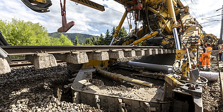 Ausschnitt einer Baumaschine auf Schienen, die ein Stück eines Gleiskörpers anhebt.