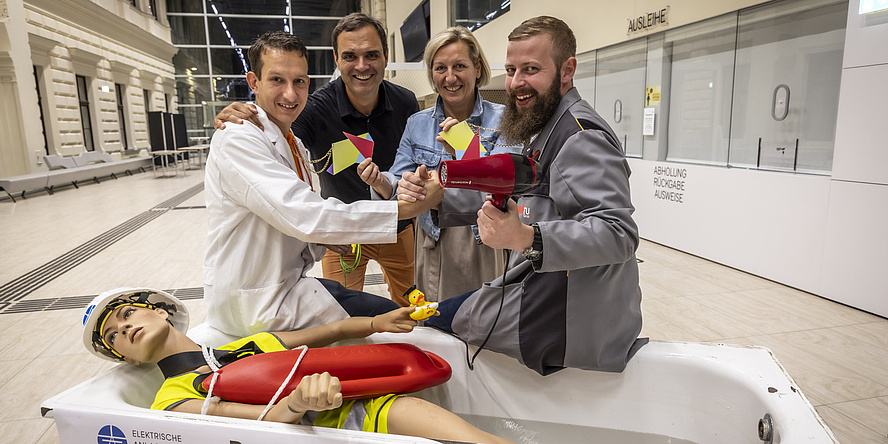 Vier Personen stehen vor einer Badewanne, in der eine Schaufensterpuppe liegt.