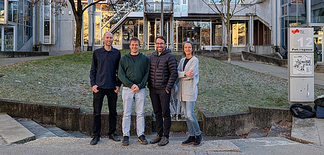Four people stand in front of a building and smile into the camera, three men and one woman.