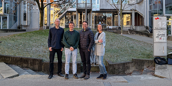 Four people stand in front of a building and smile into the camera, three men and one woman.