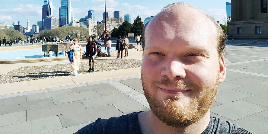 Man with bald head and full brown beard looks into the camera. He is standing outside, the sun is blinding him.