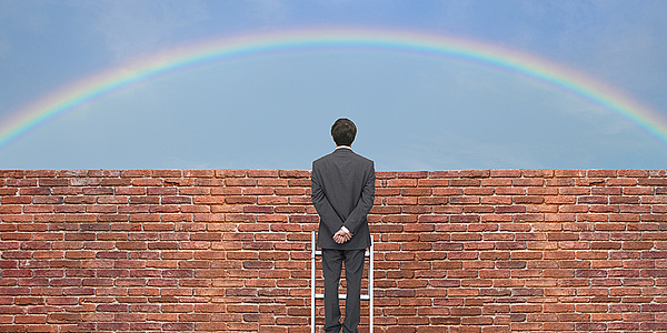 Mann steht auf Leiter und schaut über eine Mauer auf einen Regenbogen