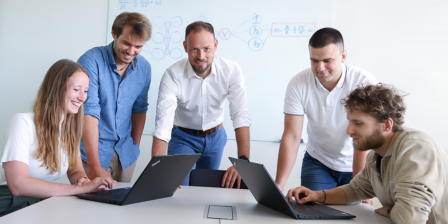 Five people are at a white table. On the far left, a woman is sitting at a laptop; on the far right, a man is sitting at a laptop. Three men are standing between them. The man in the middle is looking at the camera; the two people on the left are looking at the laptop on the left; the two people on the right are looking at the laptop on the right.