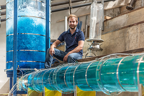 Ein junger Mann mit Bart in Jeans und blauem T-Shirt sitzt im Wasserbaulabor und ist umgeben von Versuchanordnungen in Form von wassergefüllten Plexiglas-Röhren