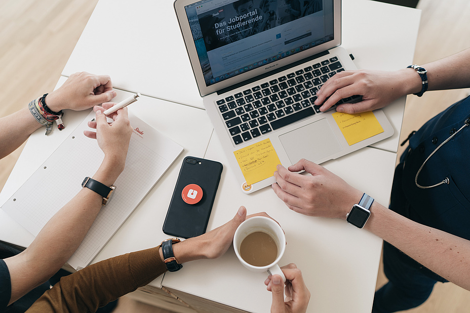 Laptop am Arbeitstisch mit drei Personen samt Mobiltelefon und Kaffeeschale