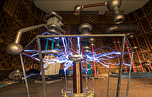 Blaue Blitze rund um eine Tesla-Spule, dahinter futuristische Objekte in einer riesigen Halle.