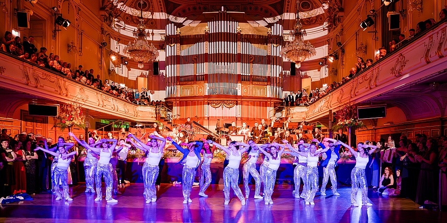 A group of young women dance lively in the Graz Congress. The ladies are a professional dance troupe. The ballroom is brightly lit. Ball guests can be seen in the background.