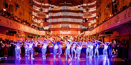 Eine Gruppe von jungen Frauen tanzt beschwingt im Grazer Congress. Bei den Damen handelt es sich um eine professionelle Tanztruppe. Der Ballsaal ist hell erleuchtet. Im Hintergrund sieht man Ballgäste.