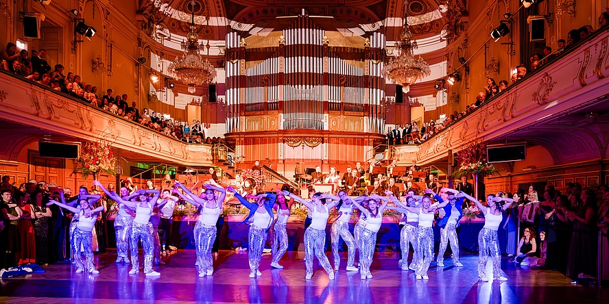 A group of young women dance lively in the Graz Congress. The ladies are a professional dance troupe. The ballroom is brightly lit. Ball guests can be seen in the background.